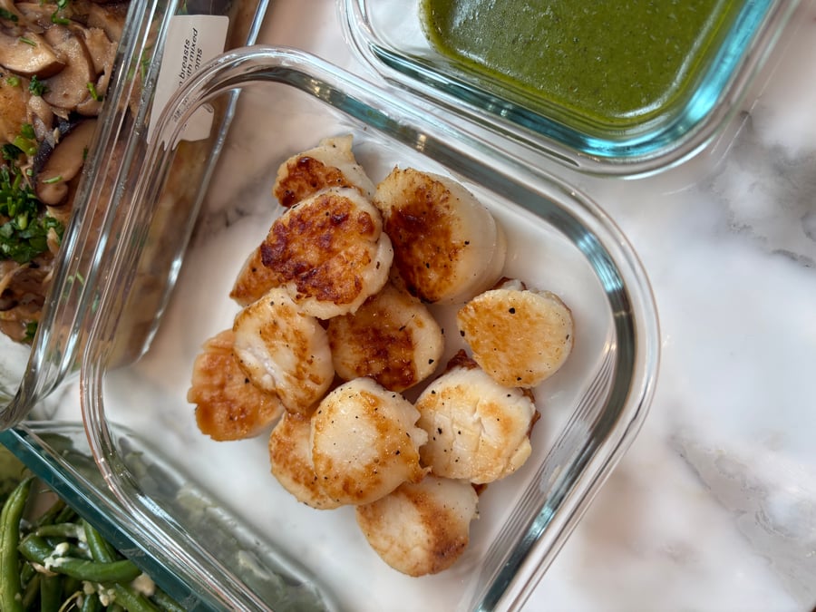 A glass container of golden seared scallops rests beside garlic goat cheese french beans and chicken breast marsala in clear meal prep dishes.