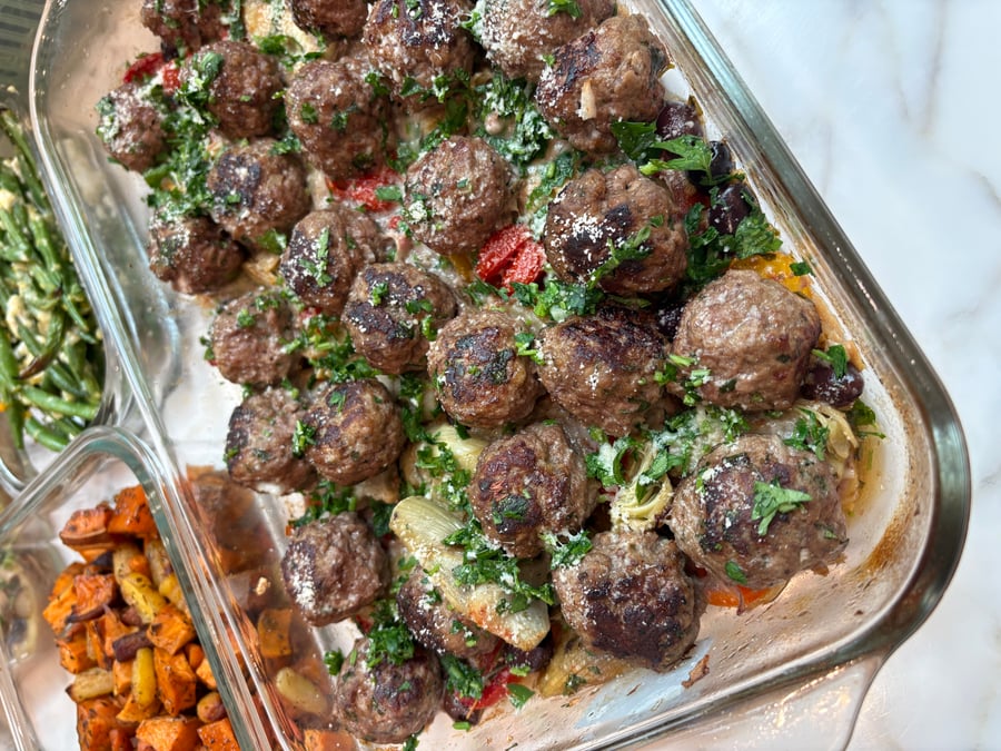 Glass meal prep containers filled with Greek beef meatballs, artichoke hearts, olives, roasted sweet potatoes and carrots, and garlic goat cheese green beans.