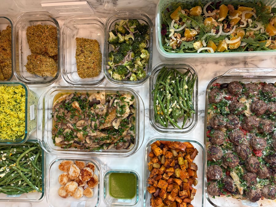 Top-down view of glass containers filled with chicken marsala, pistachio-crusted halibut, garlic scallops, greek meatballs, and assorted vegetable sides on a marble surface.