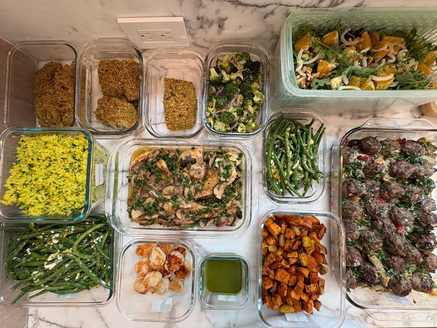 A collection of glass meal prep containers featuring chicken marsala, pistachio-crusted halibut, garlic scallops, Greek meatballs, saffron rice, roasted vegetables, and a fresh orange fennel salad.
