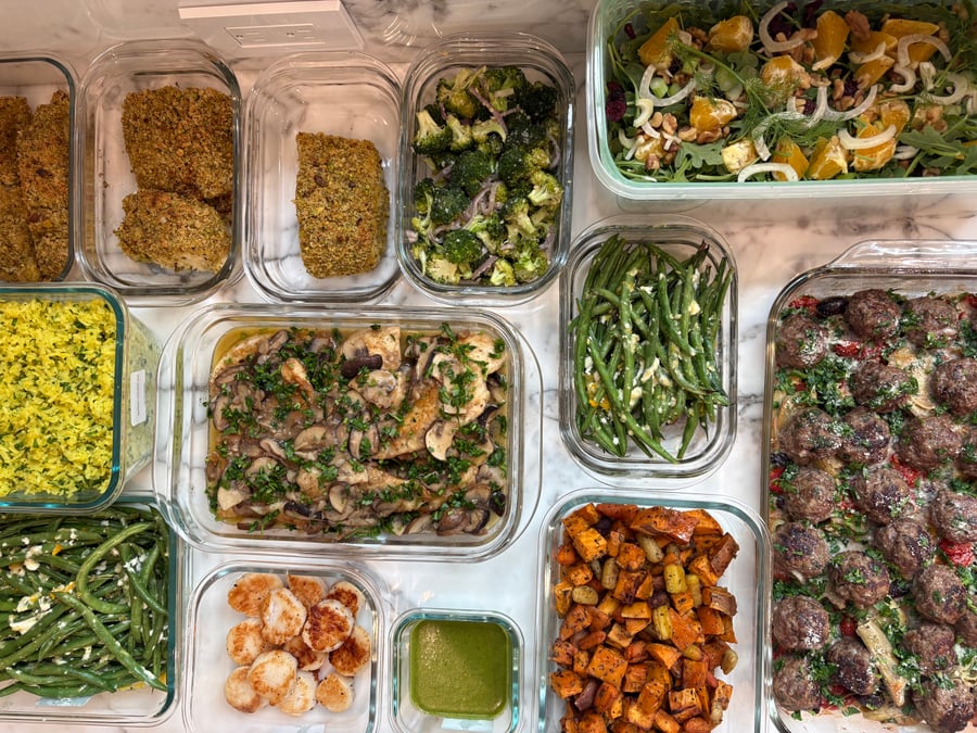 A variety of glass containers filled with pistachio halibut, chicken marsala, garlic scallops, roasted vegetables, and fresh salad arranged on a marble surface.