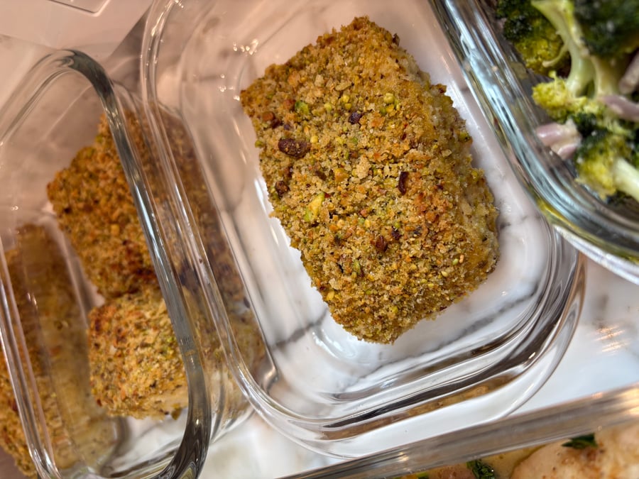 Glass containers holding golden pistachio crusted halibut fish filets, served alongside roasted miso broccoli, ready for a nutritious meal-prep service on a marble surface.