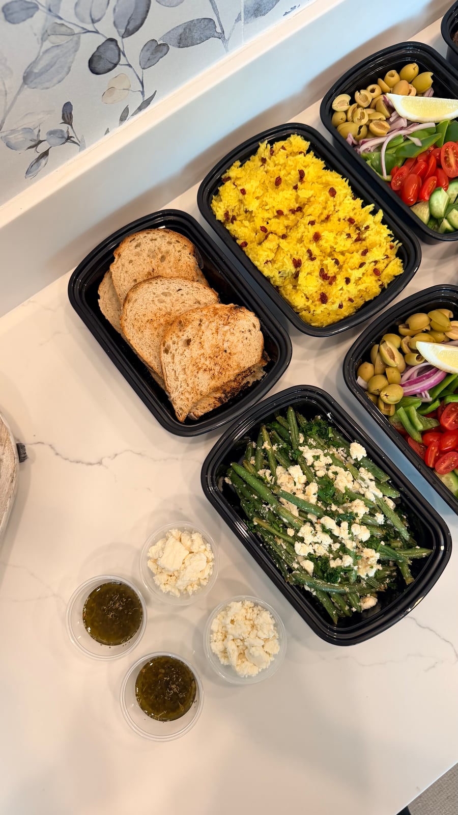 Black meal prep containers featuring sourdough bread, saffron rice, Greek salad with olives, and garlic goat cheese green beans on a white counter.