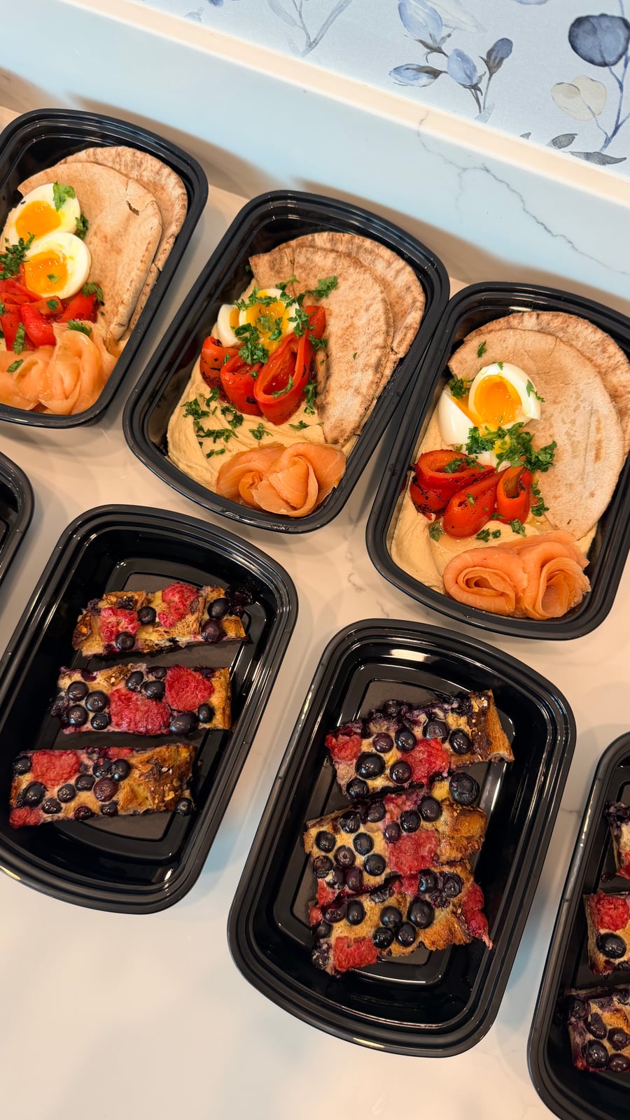 Black meal prep containers displaying hummus bowls with pita, smoked salmon, and eggs, alongside containers featuring a berry-filled french toast bake for breakfast.