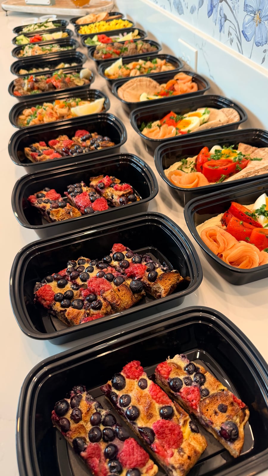 A selection of black meal prep containers featuring a protein-rich French toast bake with fresh blueberries and strawberries arranged in a tidy grid.