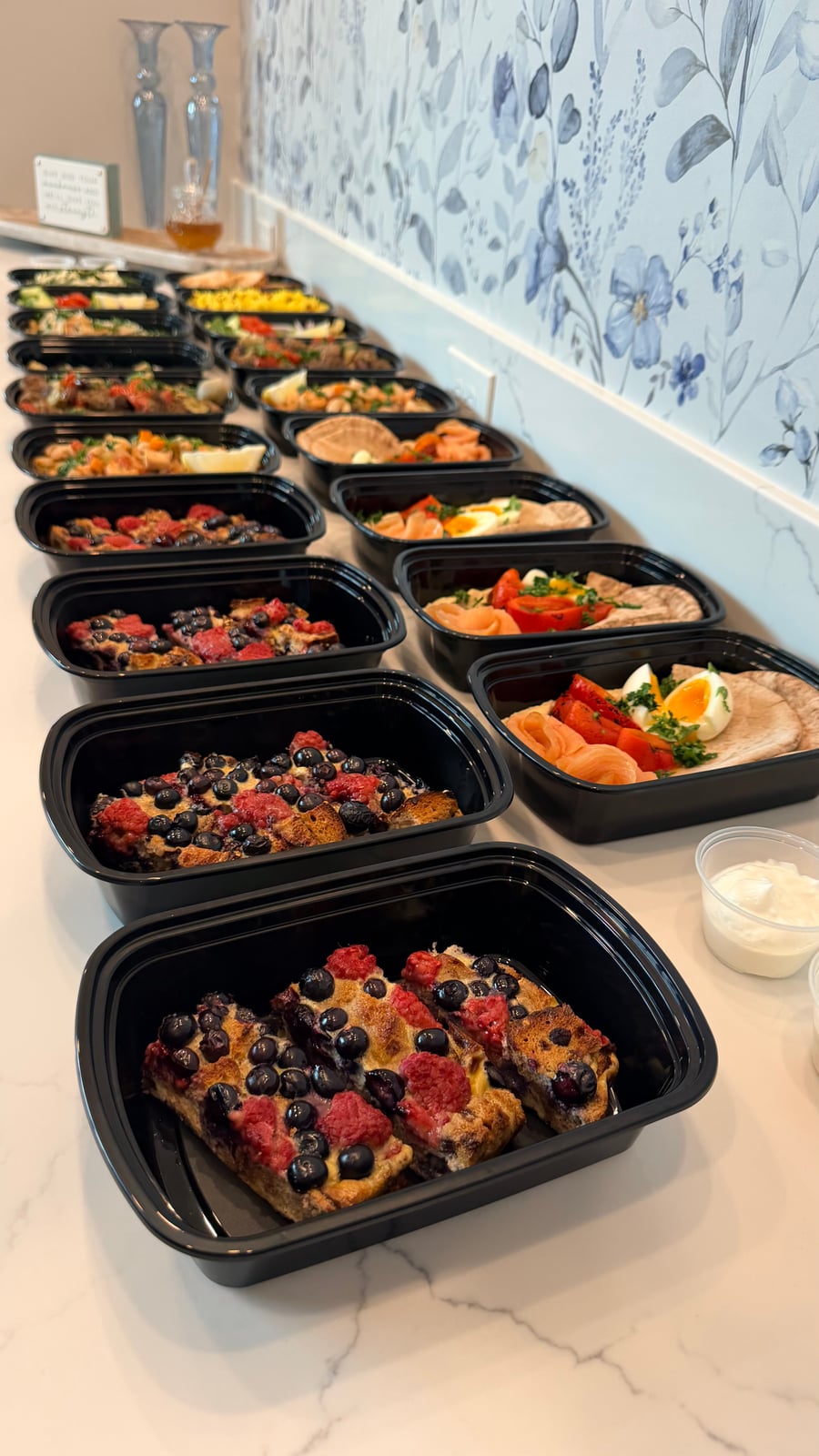 Black meal prep containers displaying breakfast portions of strawberry and blueberry French toast bake served alongside small individual pots of fresh yogurt.