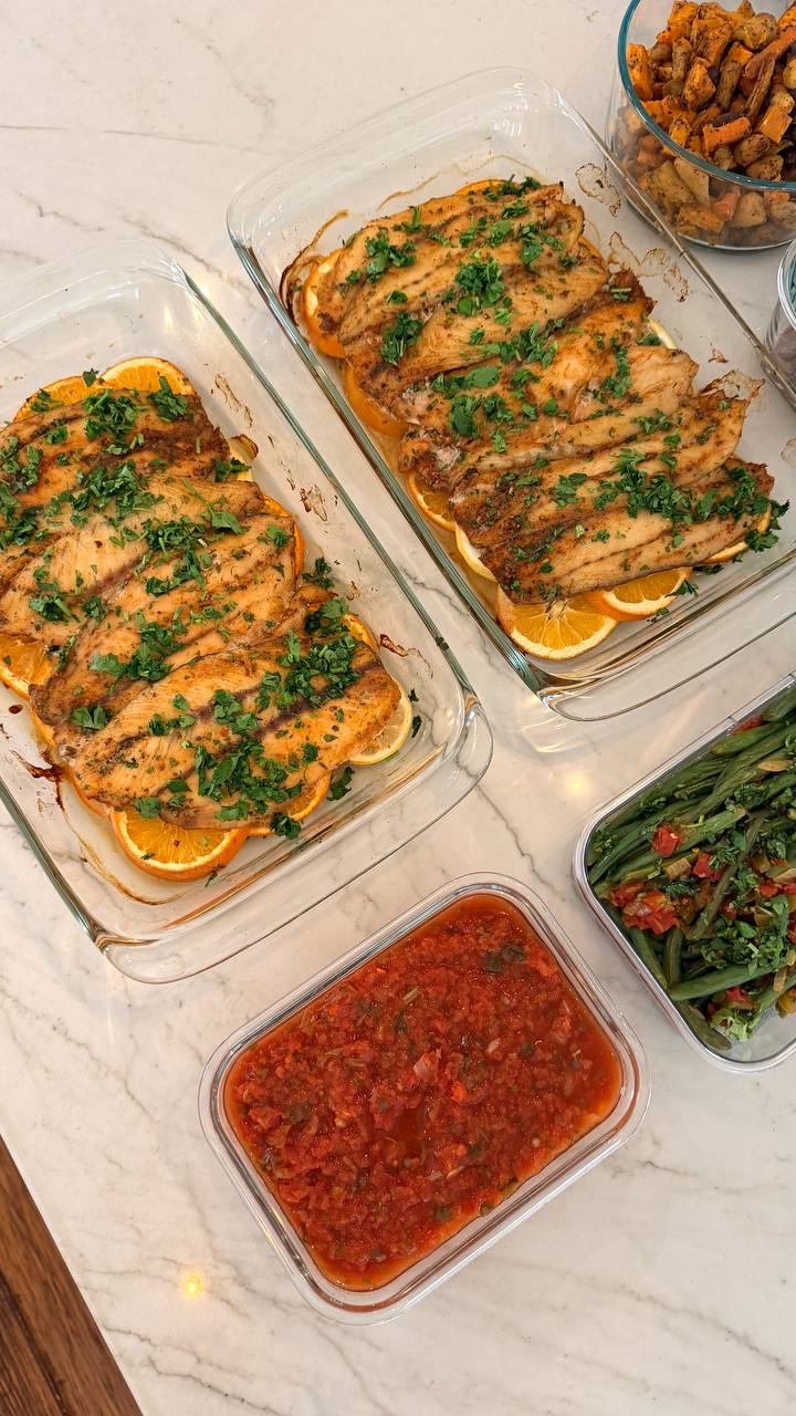 Glass baking dishes containing orange lemon barramundi, fresh tomato salsa, Mexican style green beans, and roasted sweet potatoes and carrots on a marble counter.
