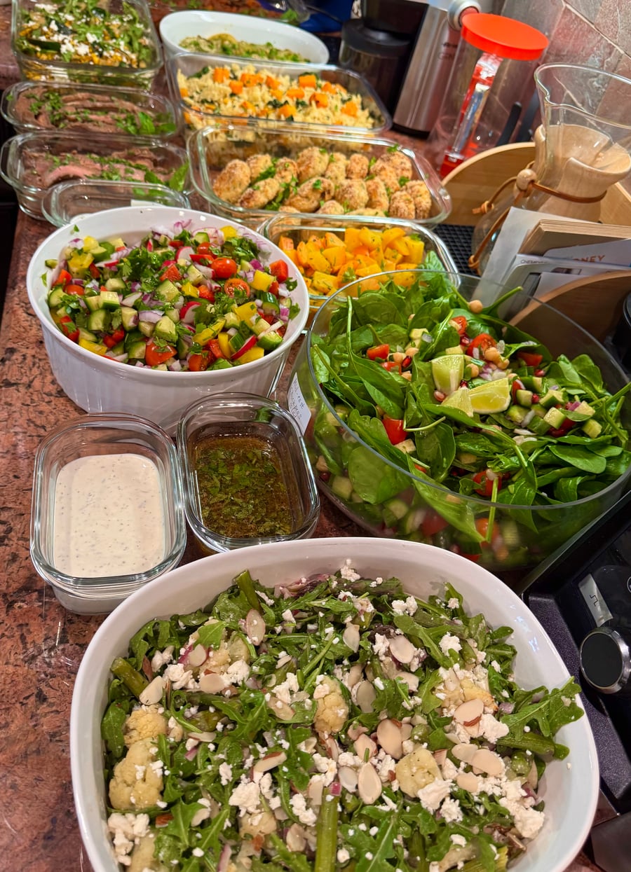 A variety of vibrant salads including arugula, cucumber-tomato, and baby spinach, alongside roasted cauliflower, butternut squash, and protein in glass serving dishes.
