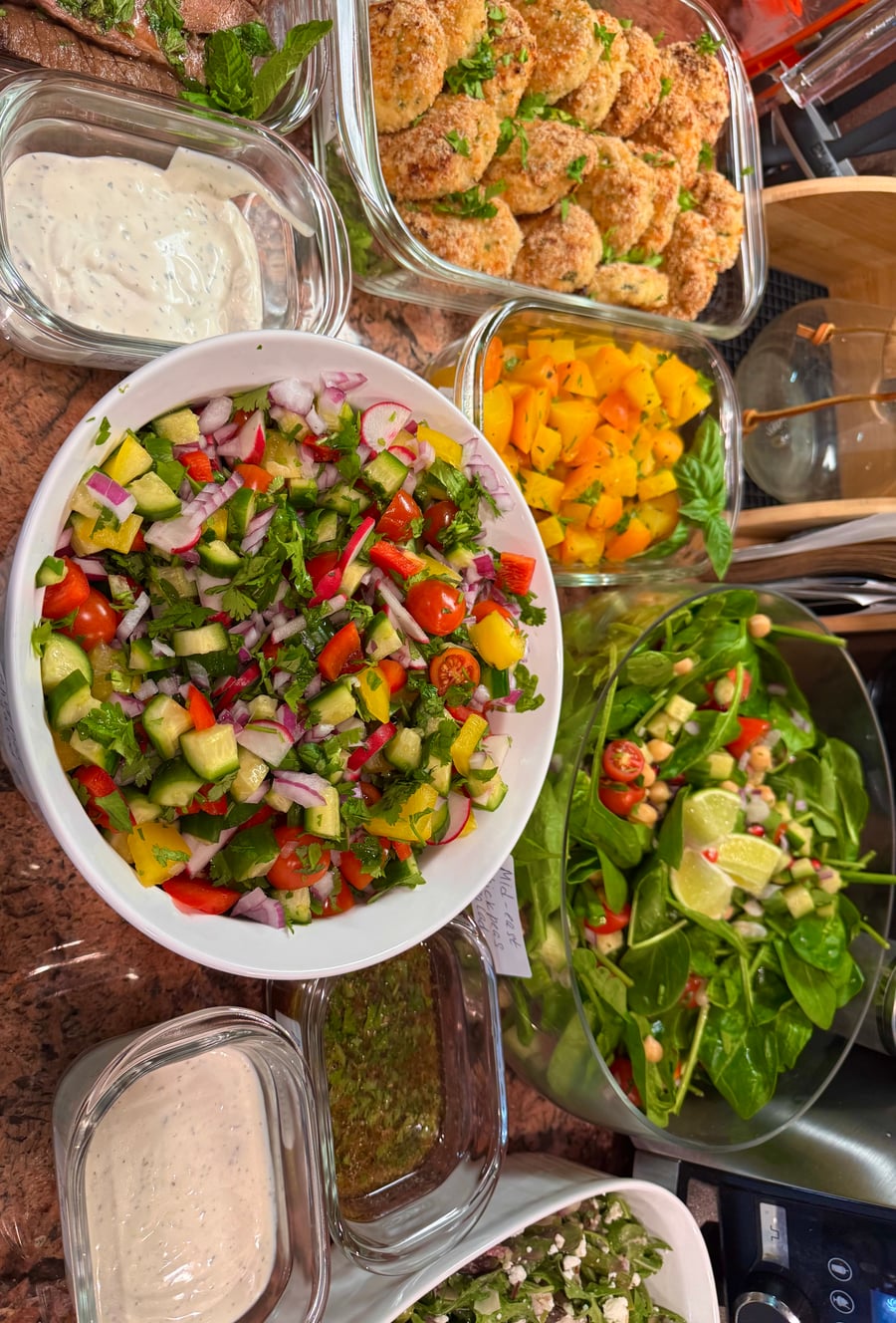 An overhead collection of glass containers featuring crispy breaded chicken bites, a vibrant chopped vegetable salad, roasted yellow squash, and a fresh spinach salad.
