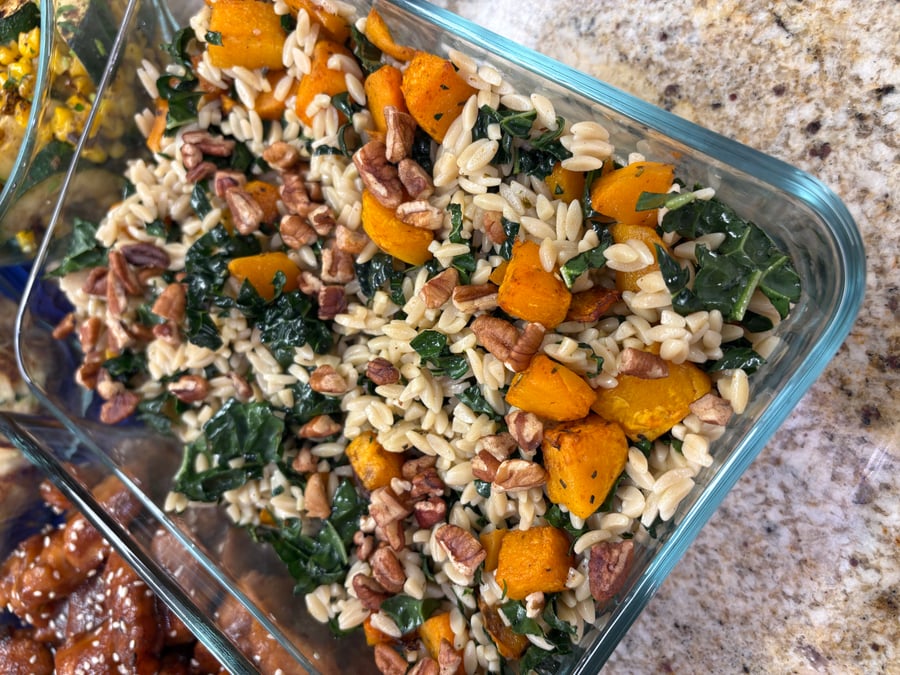 A glass meal prep container filled with tender brown rice orzo, roasted butternut squash, dark leafy kale, and crunchy toasted pecans.