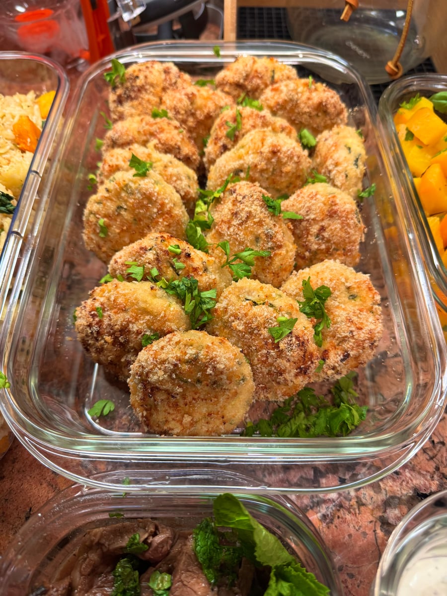 A glass rectangular container filled with golden-brown breaded potato cakes garnished with fresh green parsley on a marble countertop.