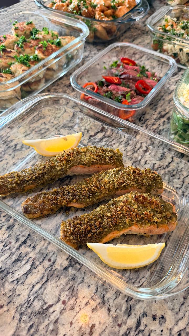 Three roasted herb-crusted salmon fillets with lemon wedges arranged neatly in a glass container for professional meal preparation.