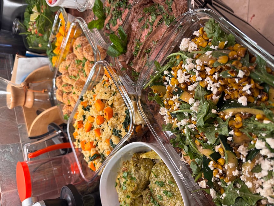 A collection of glass meal prep containers featuring pesto chicken, roasted vegetable rice, breaded meatballs, seared steak, and a corn zucchini salad.