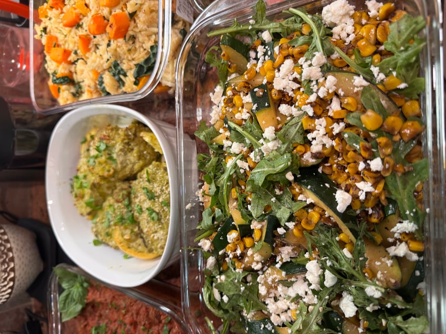 A variety of glass containers featuring pesto herb chicken, savory orzo with carrots, and a fresh zucchini corn salad topped with feta cheese and arugula.