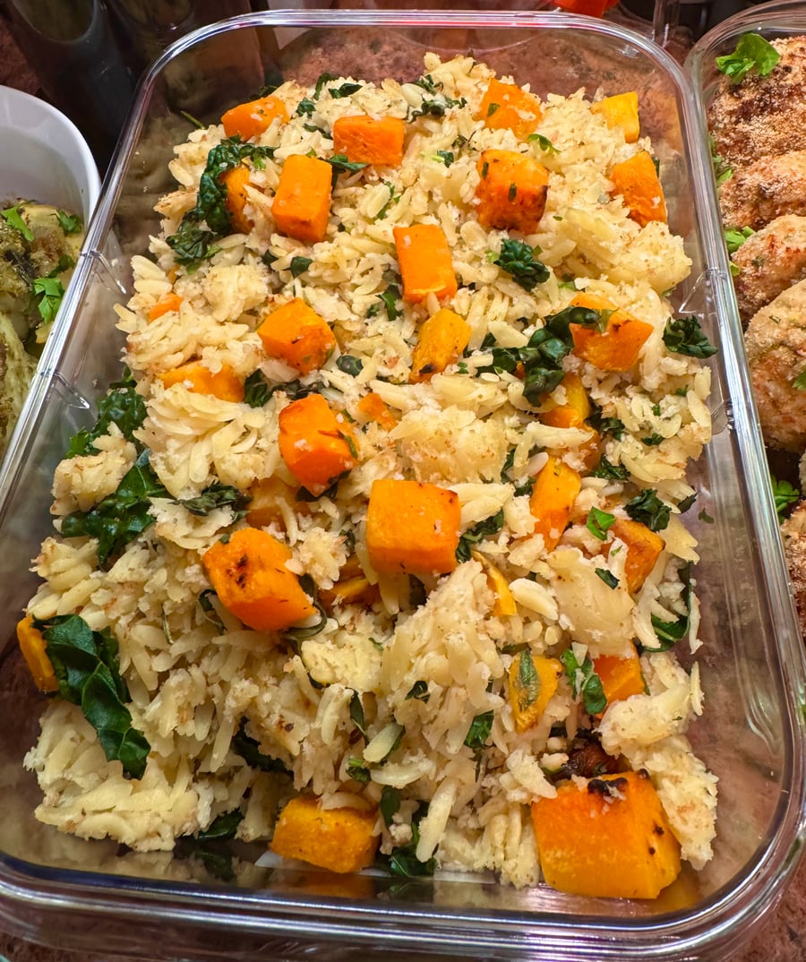 A glass tray filled with a vibrant orzo salad featuring cubes of roasted butternut squash, fresh dark kale leaves, and a light herb seasoning.
