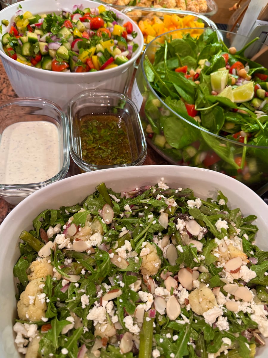 A variety of vibrant, freshly prepared salads featuring roasted cauliflower, spinach, chopped vegetable salsa, crumbly feta, and toasted almond slivers in glass bowls.