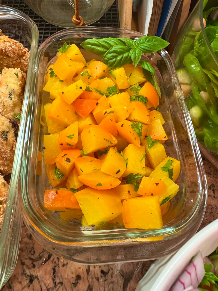 A glass container filled with vibrant roasted golden beet cubes, garnished with fresh green parsley and topped with two bright green basil leaves.
