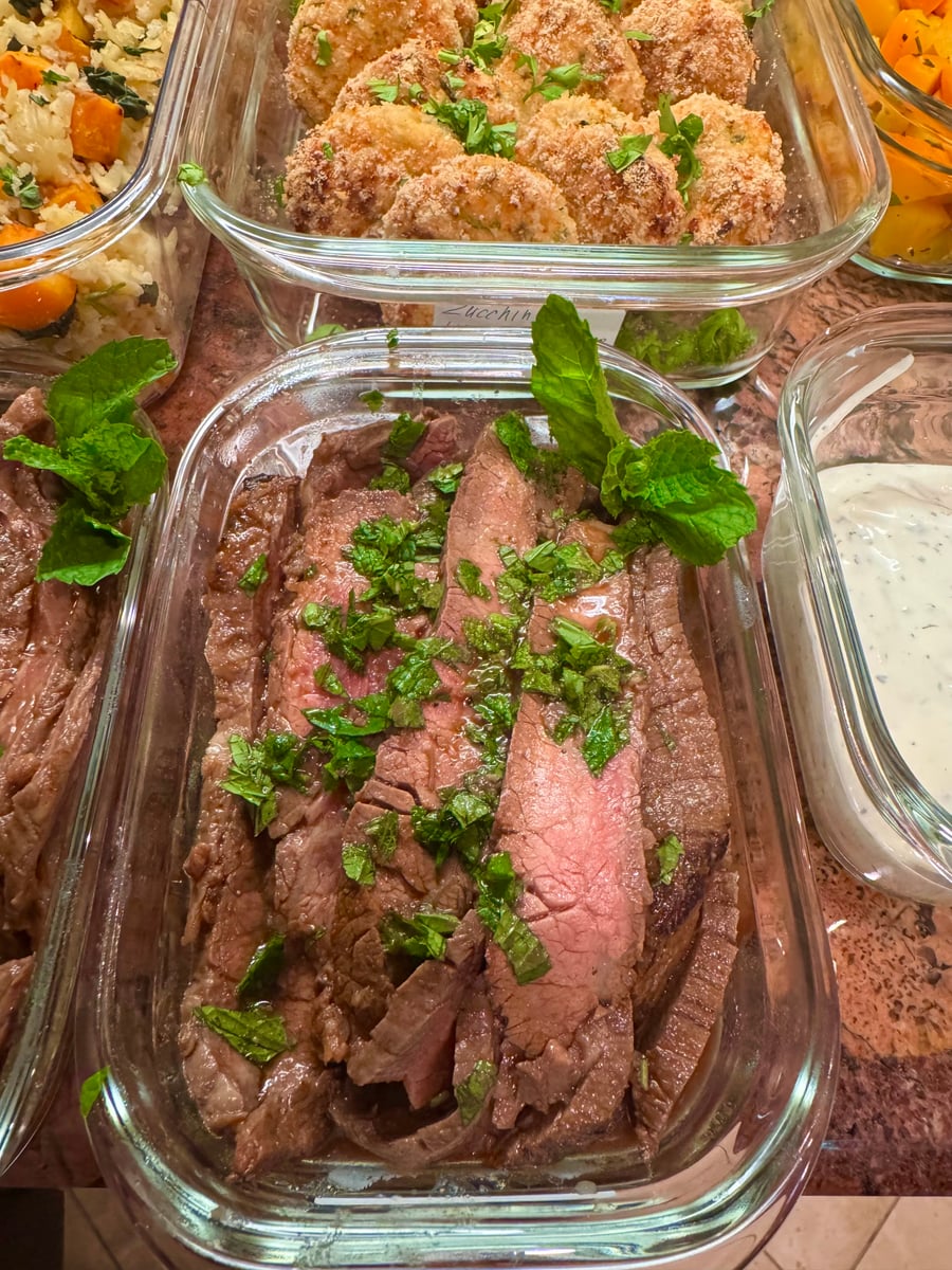 Glass meal prep container featuring tender, medium-rare sliced steak garnished with fresh chopped mint, presented alongside other containers of zucchini patties and dipping sauce.