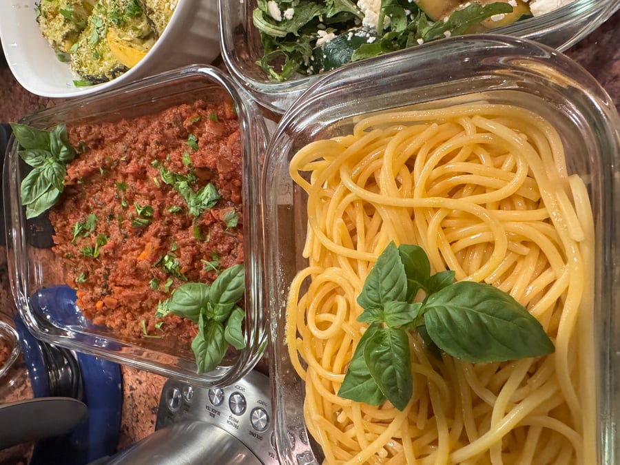 Glass containers featuring spaghetti pasta and a rich meat bolognese sauce, both garnished with fresh vibrant green basil leaves for an elegant presentation.