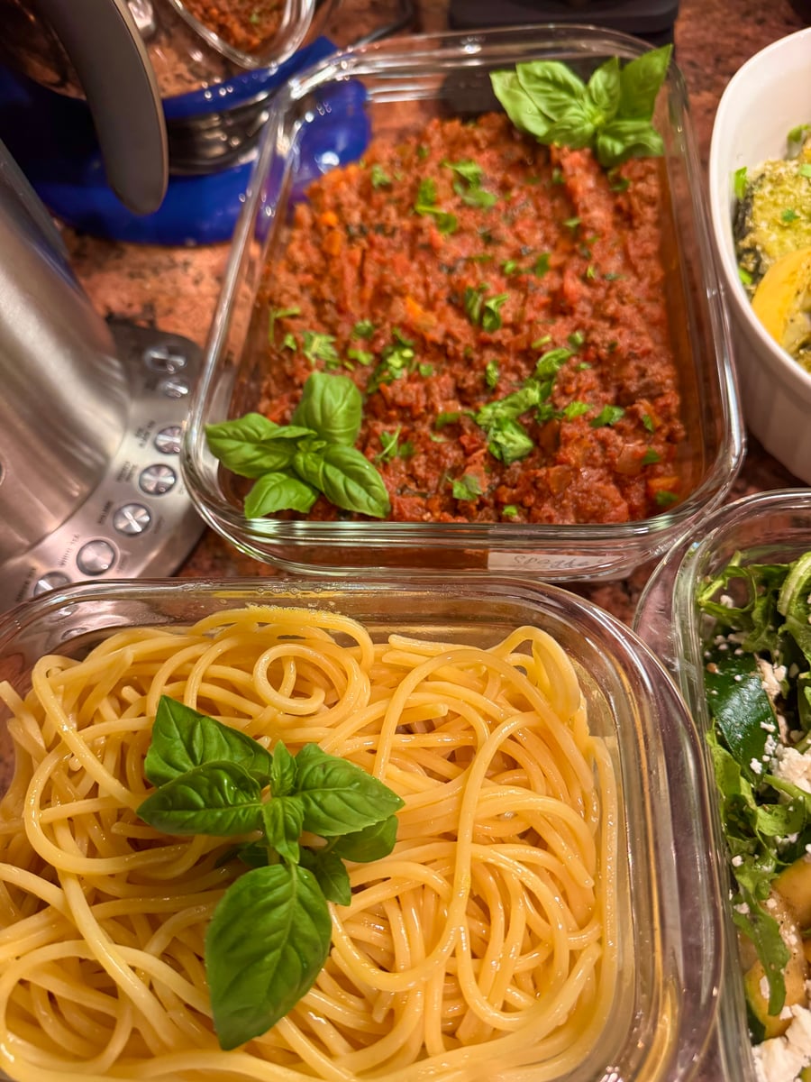 A glass container of rich, red beef bolognese sauce and a separate container of plain spaghetti, both garnished with fresh vibrant green basil leaves.