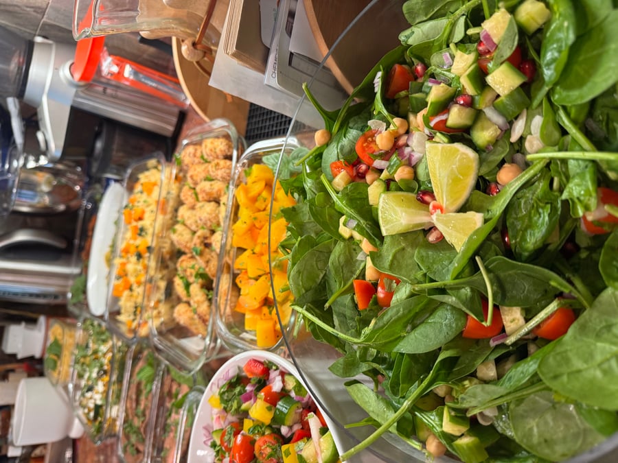 A large bowl of spinach, chickpea, and pomegranate salad next to several glass containers filled with roasted squash, breaded chicken, and colorful vegetable mixtures.