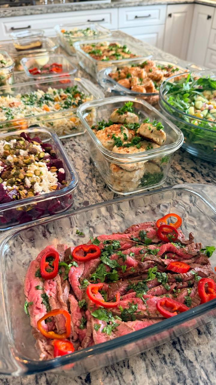 A variety of gourmet meal prep containers with steak, fritters, beets, and orzo arranged on a kitchen countertop.