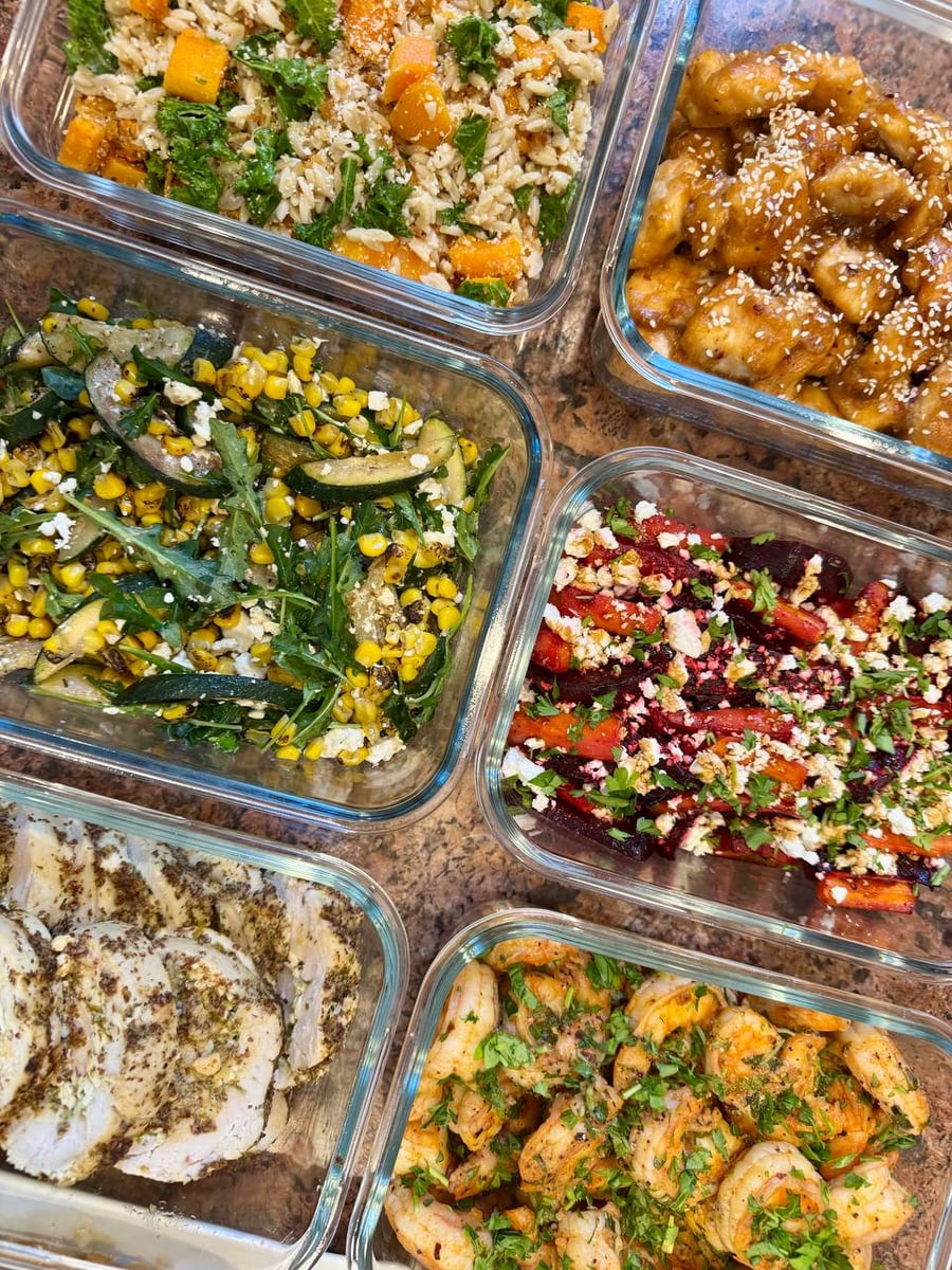 Glass containers holding feta stuffed chicken, orange chicken, shrimp, roasted beets, orzo, and charred corn vegetable salad arranged together.