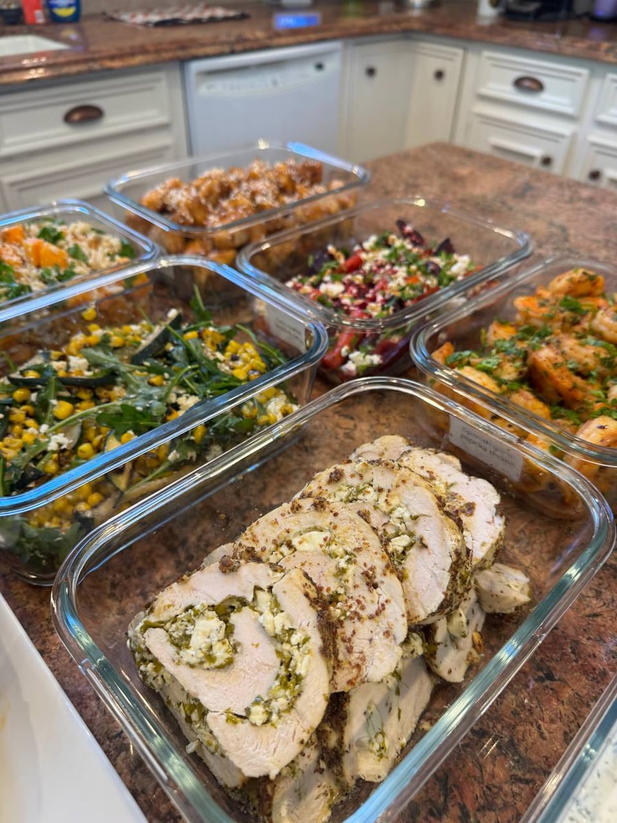 Several glass containers filled with stuffed chicken roulade, orange glazed chicken, garlic paprika shrimp, feta beets, and charred corn with zucchini side dishes.