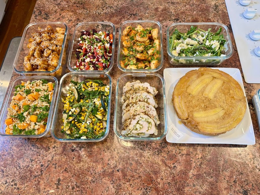 Glass meal prep containers filled with orange chicken, feta beets, garlic shrimp, and veggie sides alongside a miso maple upside-down banana cake.