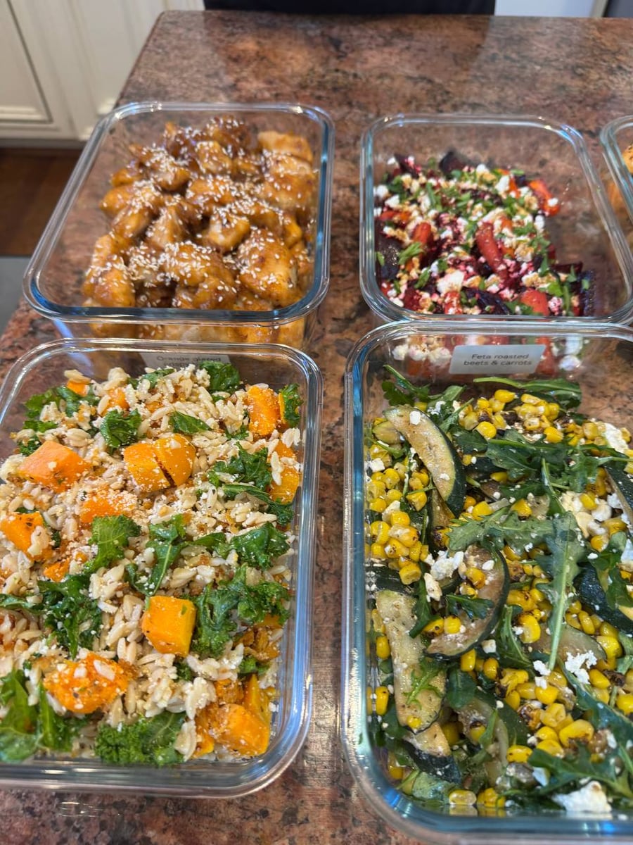 Glass containers featuring orange glazed chicken bites, feta roasted beets and carrots, butternut squash rice orzo, and charred corn with zucchini and arugula.