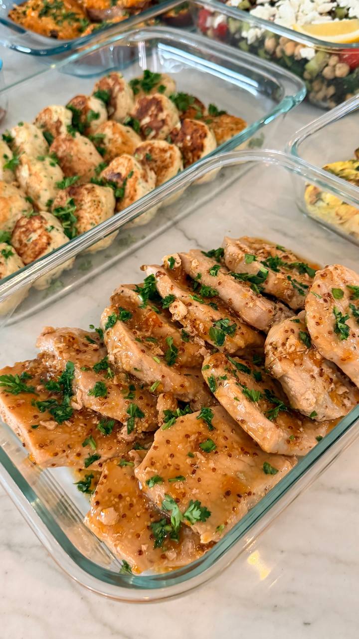 A glass container holds succulent, sliced apricot glazed pork loin chops, topped with fresh parsley and visible mustard seeds, presented on a marble surface.