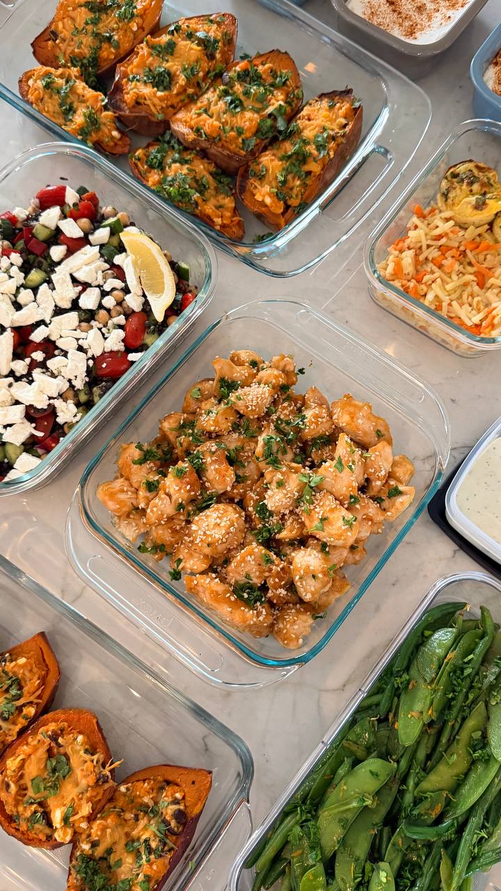 Glass containers featuring orange glazed chicken, Mediterranean chickpea salad, stuffed sweet potato boats, and garlic seared green beans and sugar peas.