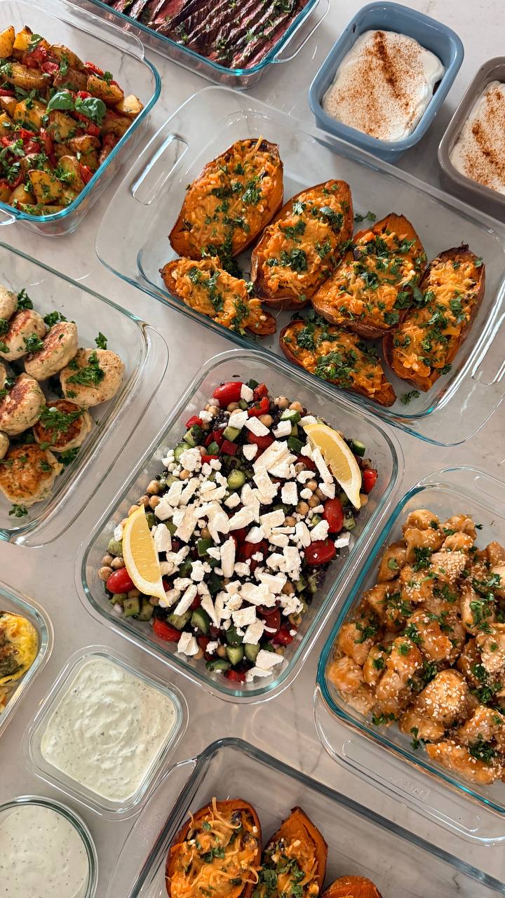 A variety of glass containers holding sweet potato boats, Mediterranean chickpea salad, orange glazed chicken, turkey fritters, roasted potatoes, and yogurt avocado sauce.