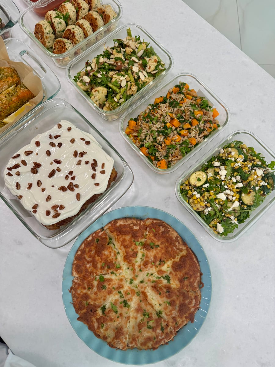 An overhead view of glass meal prep containers featuring turkey fritters, roasted cauliflower, orzo, charred corn salad, carrot cake oats, and egg tortilla pockets.