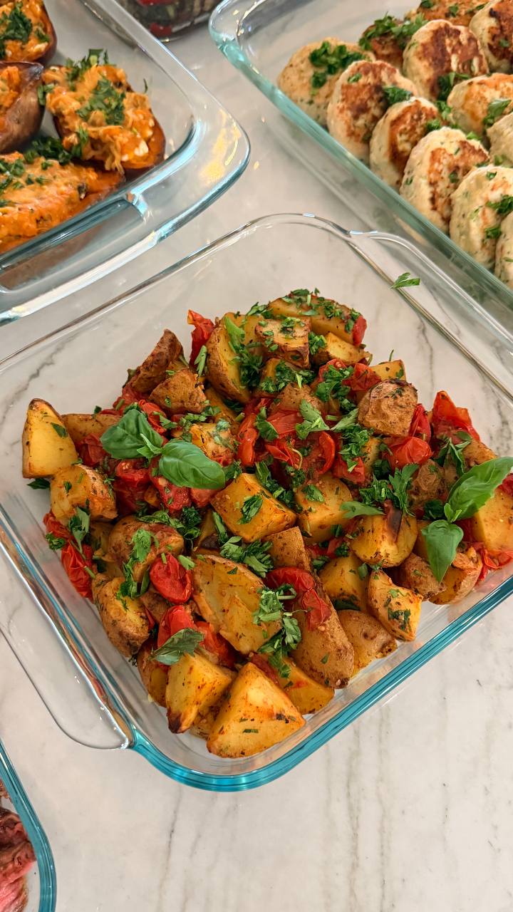 A glass dish filled with golden roasted baby potatoes and blistered cherry tomatoes, garnished with fresh green herbs and fragrant basil leaves.