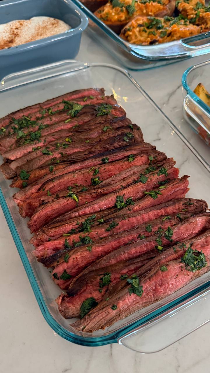 A glass container holds perfectly sliced Vietnamese flank steak garnished with fresh herbs, alongside containers featuring stuffed sweet potatoes and a creamy white sauce.