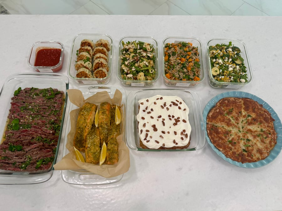An assortment of prepared dishes including sliced flank steak, pesto salmon, roasted vegetables, and baked goods arranged in glass containers on a white counter.