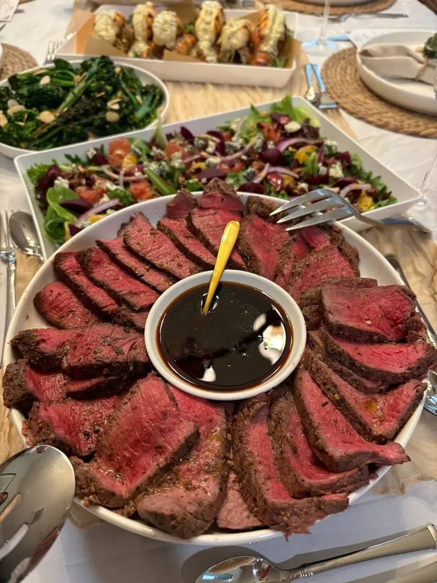 Herb-crusted beef tenderloin sliced and fanned around pomegranate sauce on a white platter, with salad and lobster in the background.