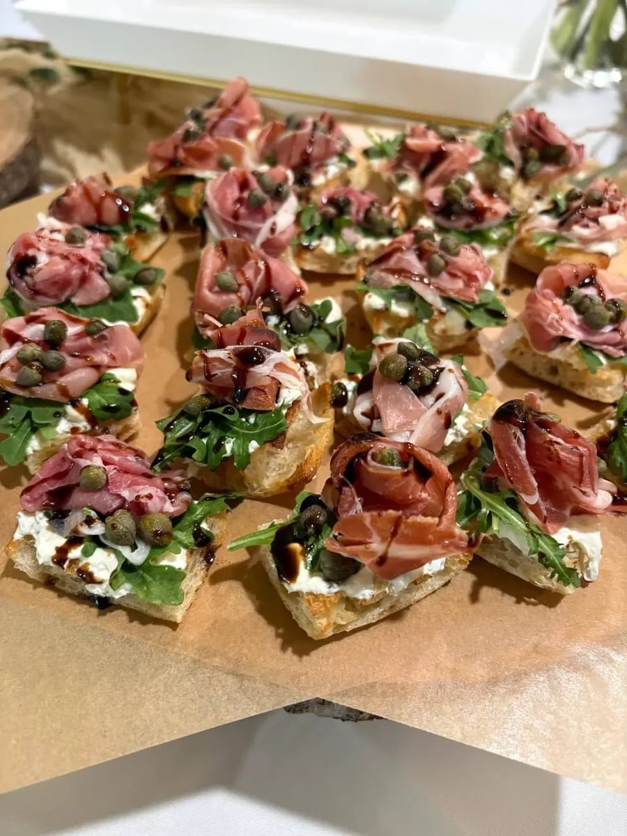 Mini prosciutto crostini with cream cheese, capers, arugula, and balsamic drizzle on toasted bread.