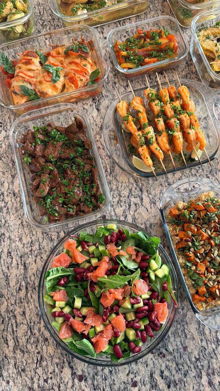 An overhead view of glass containers filled with tomato basil caprese chicken, paprika lime shrimp skewers, beef stir fry, supergreens salad, and roasted sweet potato quinoa.