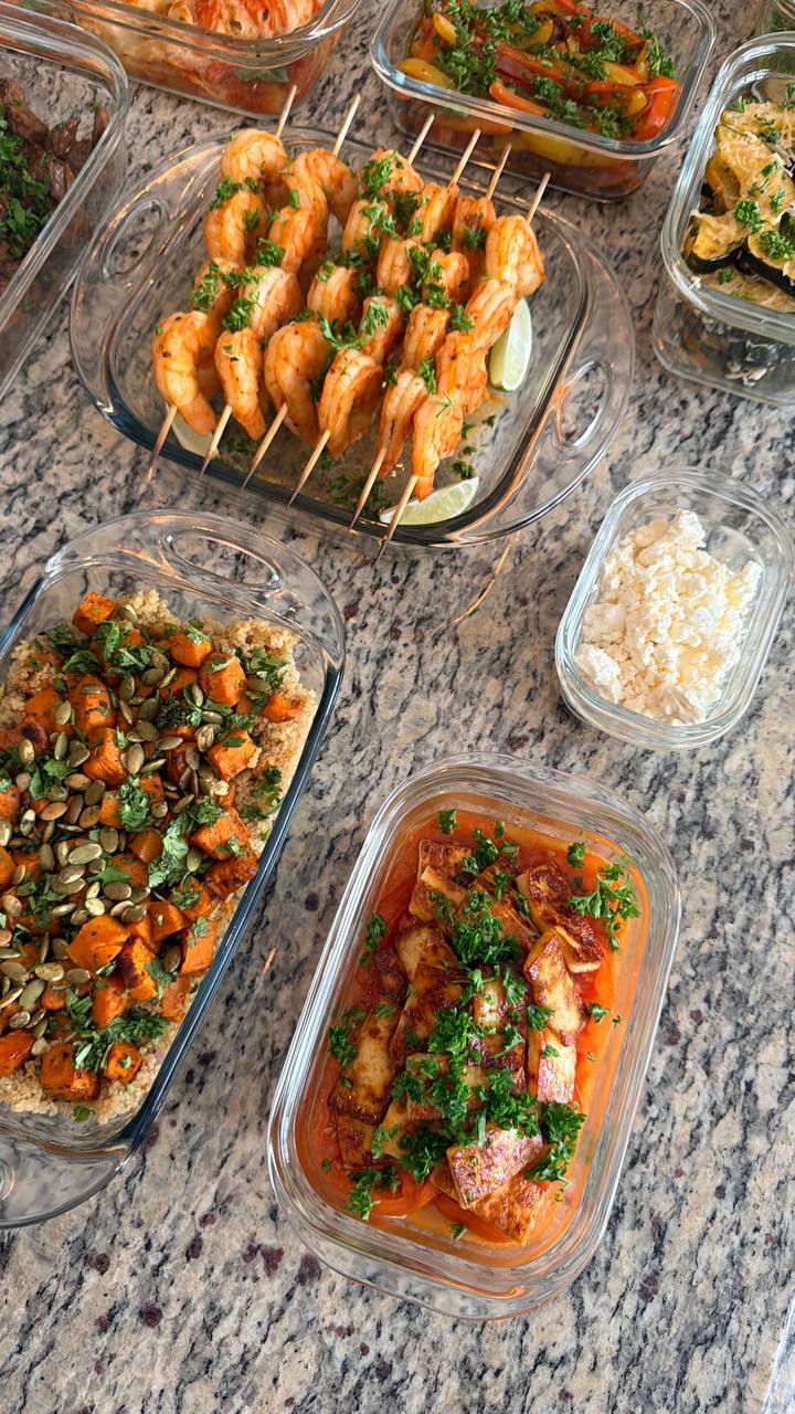 Glass meal prep containers filled with paprika lime shrimp skewers, roasted sweet potato quinoa, heirloom tomatoes with paneer, and crumbled feta cheese on granite.