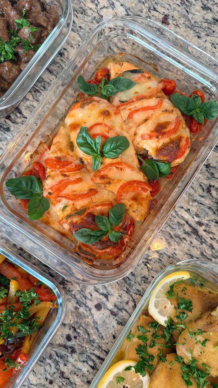 A glass serving dish containing baked tomato basil Caprese chicken breasts, topped with melted cheese, fresh tomato slices, and fragrant green basil leaves.