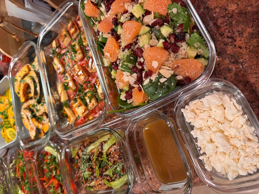 A variety of glass containers holding roasted squash, grilled halloumi with tomatoes, stir-fried broccoli, and a vibrant grapefruit spinach salad with feta cheese.