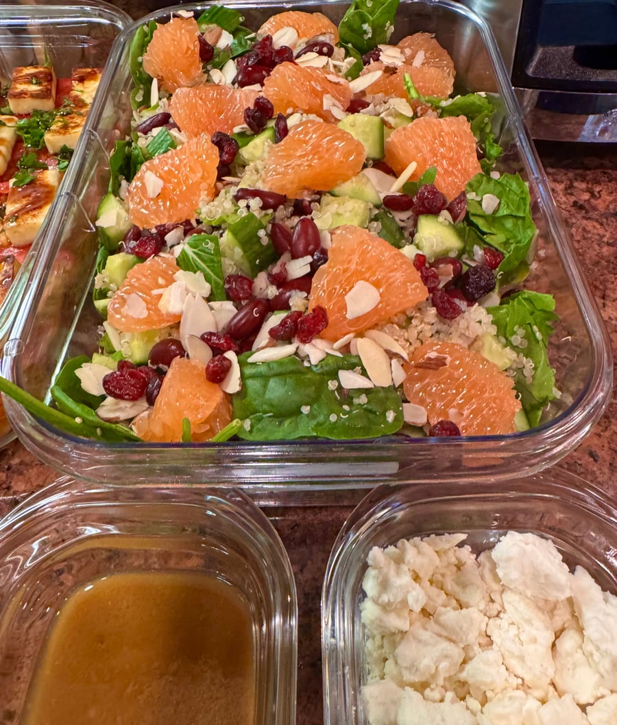 A vibrant grapefruit, quinoa, and spinach salad in a glass container, topped with sliced almonds, dried cranberries, and diced cucumbers for texture.