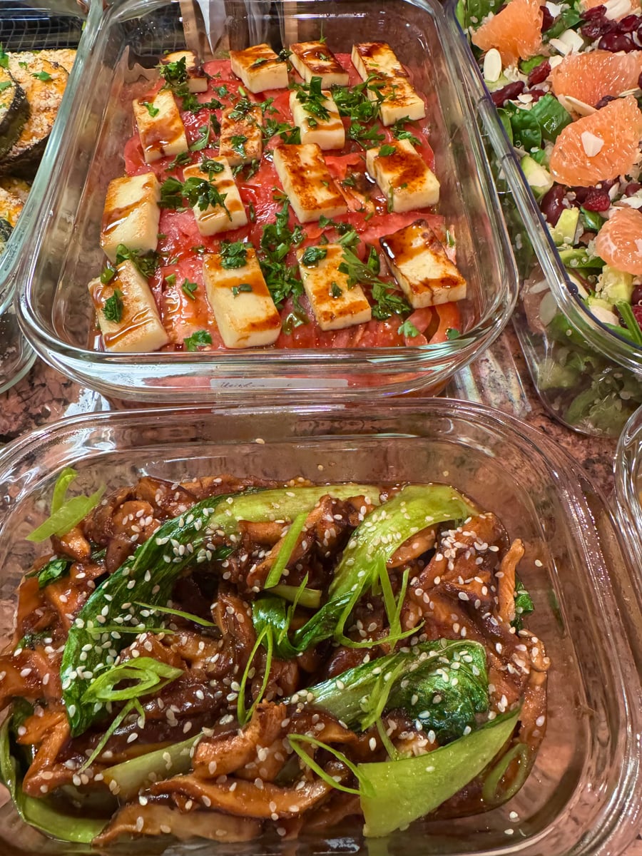 A glass meal prep container features grilled halloumi cheese over watermelon slices, while a second container holds glistening bok choy and mushroom stir-fry.