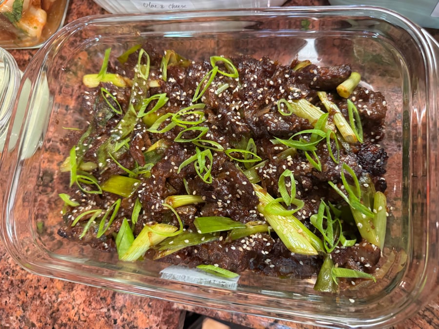 A glass container holding savory glazed beef stir-fry topped with fresh sliced green onions and toasted sesame seeds on a granite countertop surface.