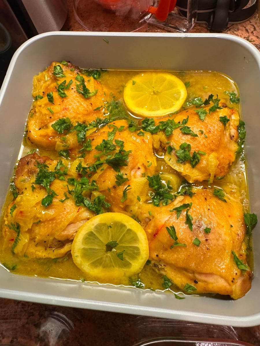 A square baking dish containing golden braised chicken thighs, bright slices of lemon, and fresh chopped parsley in a savory, aromatic pan sauce.