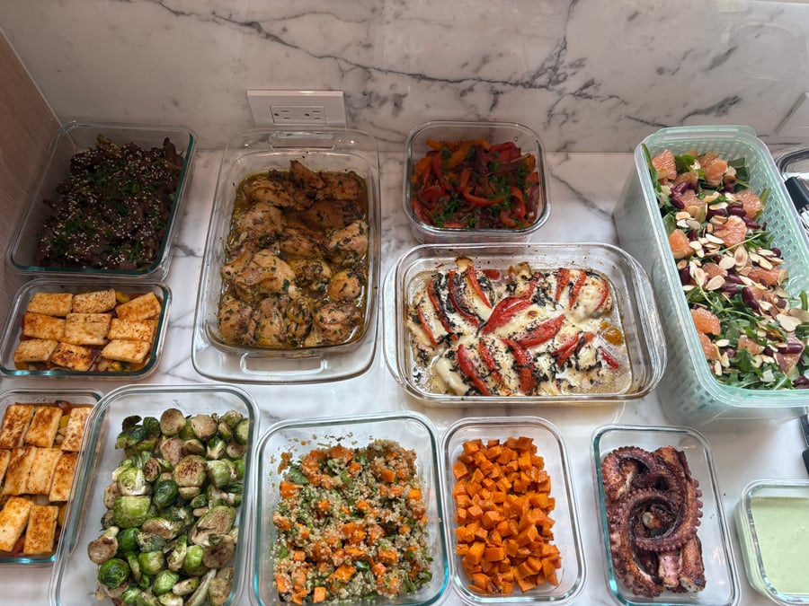 A variety of glass containers holding saffron chicken, tomato basil breasts, octopus, masala sprouts, roasted sweet potatoes, and a grapefruit supergreens salad.