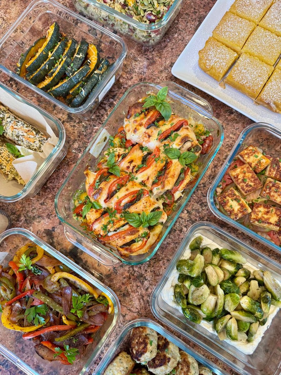 A variety of glass containers filled with tomato basil chicken, parmesan acorn squash, maple brussel sprouts, heirloom tomatoes with paneer, and lemon bars.