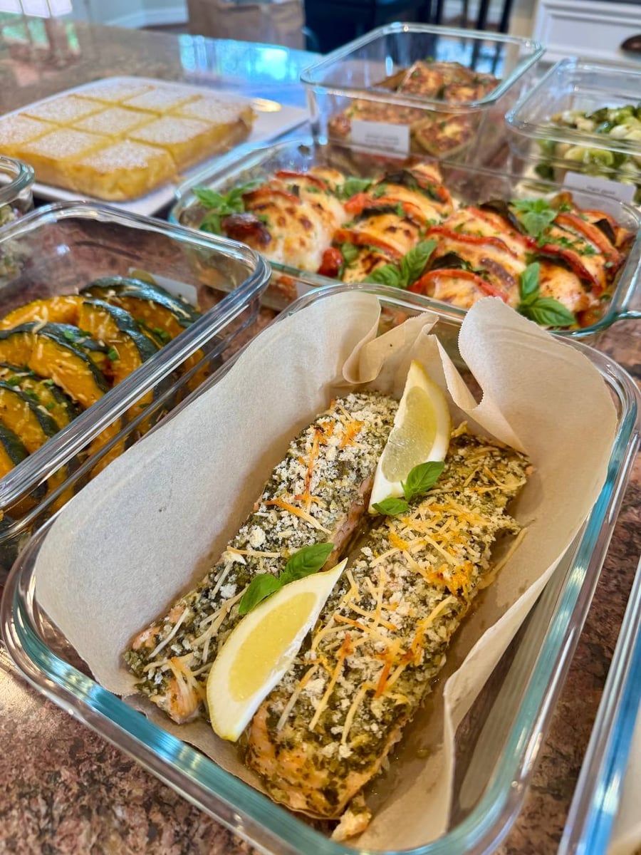 Glass meal prep containers filled with pesto crusted salmon fillets, topped with lemon wedges and fresh basil on parchment paper liners.